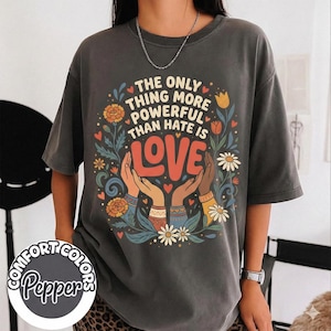 May include: Dark gray Comfort Colors t-shirt with the text "The only thing more powerful than hate is LOVE." The design includes hands, flowers, and hearts in a colorful floral arrangement. The shirt has short sleeves.