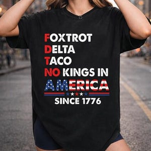 May include: Black t-shirt with the text "Foxtrot Delta Taco No Kings in America Since 1776" in red, white, and blue. The design incorporates the American flag. The shirt has a V-neck.