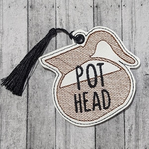 May include: A brown and white embroidered coffee pot shaped bookmark with a black tassel. The bookmark says "POT HEAD" in black embroidery.