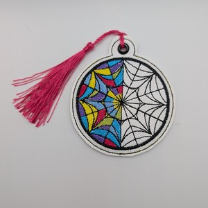 May include: Embroidered bookmark with a stained glass design. The circular bookmark features a half-colored, half-outlined design in shades of blue, yellow, purple, and red. A pink tassel is attached to the top. The bookmark is white with a black border.