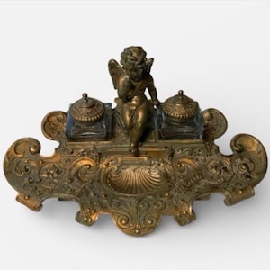 May include: An antique bronze inkwell set with a cherub figure. The inkwell has two glass ink pots with ornate lids, set on a detailed, scrolled base. A classic desk accessory, with a decorative design.