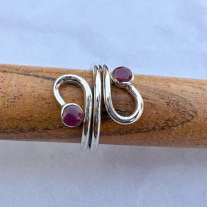 Peut inclure: Une bague en argent au design unique, ornée de deux pierres précieuses ovales rouge foncé. La bague s'enroule autour du doigt avec un motif tourbillonnant, créant un look élégant et moderne. Les pierres sont serties dans une bande d'argent poli.