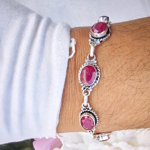 Puede incluir: Una pulsera de plata con tres piedras preciosas rojas oscuras de forma ovalada. Las gemas están engastadas en marcos de plata retorcidos y ornamentados, conectadas por delicados eslabones. La pulsera se muestra en una muñeca, mostrando el diseño y el color.