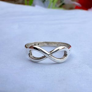 May include: A silver ring featuring an infinity symbol design. The ring is made of polished metal and is displayed on a light background. The infinity symbol is a continuous loop, representing eternity.