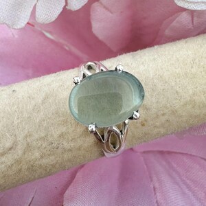 May include: A silver ring featuring a smooth, oval-shaped, pale green cabochon gemstone set in a four-prong setting. The band of the ring displays an open, decorative metalwork design that connects to the central stone setting.