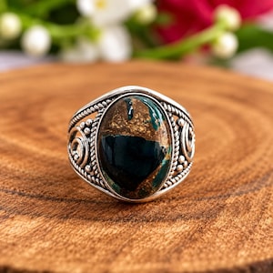 May include: A sterling silver ring featuring an oval-shaped stone with a dark blue and metallic copper-colored surface. The band is crafted with a twisted rope texture and decorative silver scrollwork accents surrounding the central gemstone.