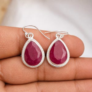 May include: A pair of teardrop-shaped earrings featuring faceted deep pink stones set in polished silver bezels. Each earring is suspended from a simple silver hook wire, showcasing a smooth, metallic border around the vibrant, textured gemstone surface.