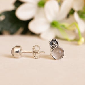 Op de afbeelding: Een paar sterling zilveren oorknopjes met een ronde, doorschijnende cabochonsteen gezet in een gepolijste metalen zetting. Het ontwerp bevat een klein, verhoogd cirkelvormig accent met drie kleine zilveren bolletjes boven de steen.
