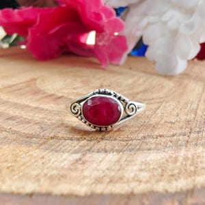 May include: A silver ring featuring a central oval-shaped faceted deep red gemstone. The metal band displays intricate scrollwork and small bead-like details surrounding the stone setting, creating a textured appearance on the silver surface.