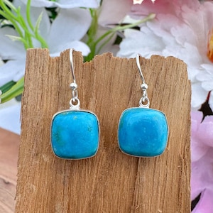 Puede incluir: Un par de pendientes colgantes con piedras turquesas cuadradas de bordes suaves y redondeados. Cada piedra está engastada en un marco de metal plateado y cuelga de un gancho sencillo de metal plateado, creando un diseño limpio y equilibrado.