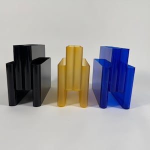 Könnte beinhalten: Drei moderne, geometrische Vasen in Schwarz, Bernstein und Blau. Die Vasen haben eine rechteckige Form mit vertikalen und abgewinkelten Elementen, die ein einzigartiges architektonisches Design schaffen. Die Vasen bestehen aus glänzendem Material.