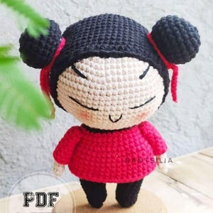 May include: A handmade crochet doll with a black hair bun, black hat, and red outfit. The doll has a smiling face and is standing on a small wooden base. The image includes the text "PDF PATTERN".