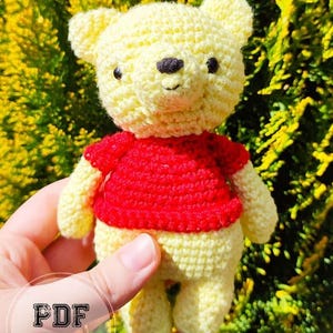 Könnte beinhalten: Ein handgehäkelter Bär in Gelb und Rot. Der Bär hat einen gelben Körper und Kopf, mit einem roten Shirt. Das Gesicht des Bären hat schwarze Augen und eine schwarze Nase. Der Text "PDF PATTERN" ist im Bild zu sehen.