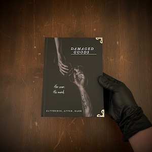 May include: A book with a dark cover featuring the text "DAMAGED GOODS" and the phrases "Her scar. His mark." The cover art depicts two hands, one reaching out to the other. Gold corner protectors are visible. The book is held by a gloved hand.