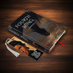May include: A hardcover book titled "Fourth Wing" with a dragon illustration and a bookmark. The bookmark features a portrait and a quote. The book has a red ribbon. The items are on a wooden surface.