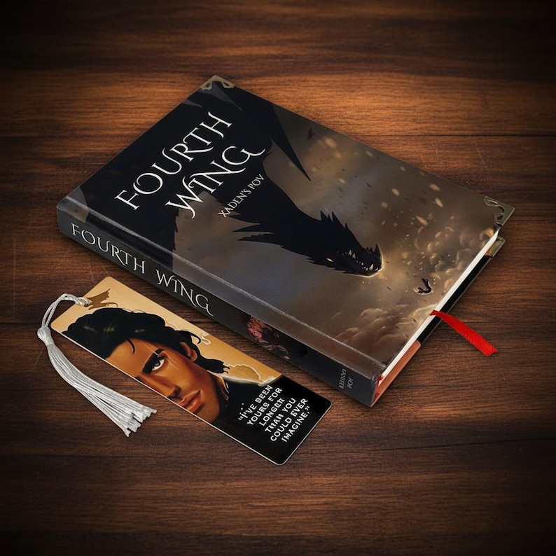 May include: A hardcover book titled "Fourth Wing" with a dragon illustration and a bookmark. The bookmark features a portrait and a quote. The book has a red ribbon. The items are on a wooden surface.