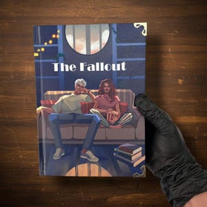 May include: A book titled "The Fallout" with a cover illustration of two people on a couch. The cover art features a blue background with a window, string lights, and a stack of books. The book has gold corner protectors.