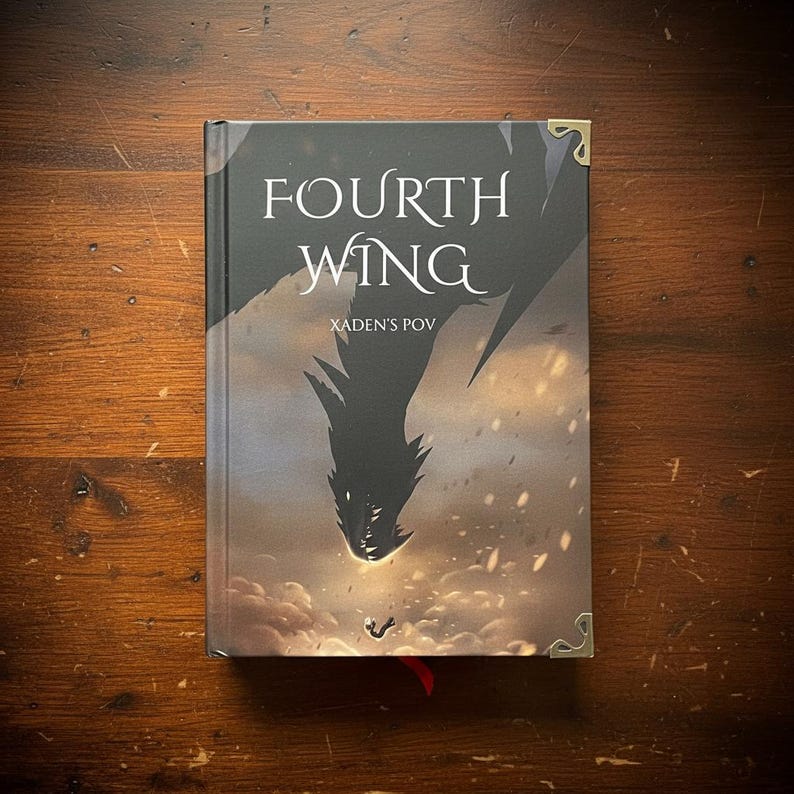 May include: A hardcover book titled "Fourth Wing" with "Xaden's POV" below the title. The cover features a dark dragon silhouette against a cloudy, fiery background. Gold-colored corner protectors and a red ribbon bookmark are visible.