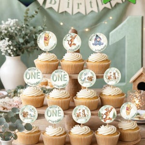 May include: A set of twelve cupcake toppers featuring Winnie the Pooh characters, woodland animals, and text including Bella's ONE 1st birthday, Thank you for coming, and Welcome. The circular paper toppers are attached to wooden picks.