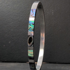 May include: A silver-toned bangle bracelet featuring alternating rectangular segments of polished metal and iridescent abalone shell inlay. The bracelet has a smooth, reflective finish and a consistent width, showcasing a repeating pattern of colourful shell.