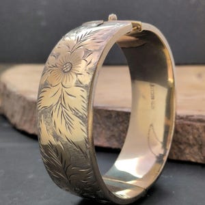 May include: A gold-toned hinged bangle bracelet featuring an intricate engraved floral pattern on the outer surface. The bracelet has a polished interior finish and a secure clasp mechanism at the top, showcasing detailed leaf and petal motifs.