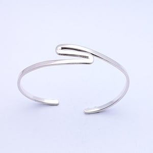 May include: A silver cuff bracelet with an open design. The bracelet has a curved, wave-like shape in the centre. The ends of the bracelet are open, allowing it to be slipped onto the wrist. The metal has a polished, reflective surface.