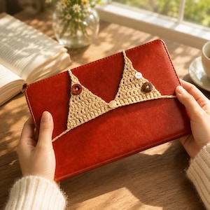 Peut inclure: Une housse de tablette en cuir rouge avec un motif de haut de bikini en crochet beige. Le haut en crochet comporte deux boutons marron et deux boutons blancs. La housse est tenue par les mains d'une personne.