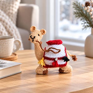 Könnte beinhalten: Eine gehäkelte Kamelfigur in Beige mit einer weiß-roten Satteldecke, die einen kleinen silbernen Akzent, eine gelbe Quaste und einen schwarzen Schwanz aufweist. Das Kamel hat dunkel gestickte Augen und einen gestickten Mund.