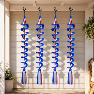 May include: Four identical spiral-shaped crochet wind spinners hanging from black hooks. Each spinner features a repeating pattern of blue, white, and red yarn, ending with a long, multi-colored tassel in blue, white, and red at the bottom.