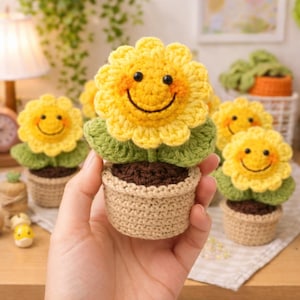 以下が含まれることがあります： 笑顔の顔、緑の葉、茶色の中心部を持つ、かぎ針編みの黄色のヒマワリ。ベージュのかぎ針編みの鉢に植えられています。手作りの花はオレンジ色の頬と黒い目があります。陽気で気まぐれなクラフトプロジェクト。