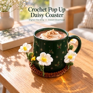 May include: A crochet pop up daisy coaster featuring white flowers with yellow centres and green stems, surrounding a dark green mug with a holly and snowflake pattern. The text reads Crochet Pop Up Daisy Coaster, Digital File Only, Instant Download.