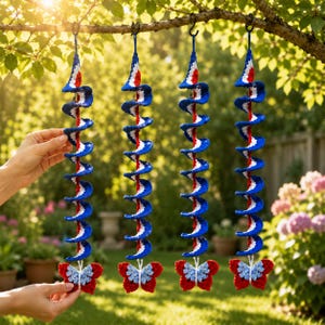 May include: Four hanging crochet spiral decorations in red, white, and blue, each featuring a butterfly at the bottom. The spirals are constructed with a central red cord and blue and white yarn, suspended from black hooks on a tree branch.