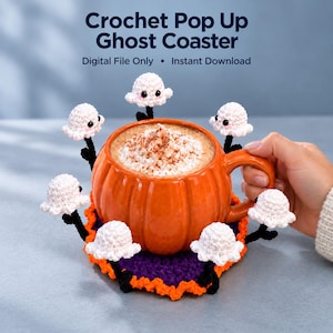Op de afbeelding: Een ronde, paars en oranje gehaakte onderzetter met zes kleine, witte, spookvormige figuren met zwarte steeltjes en roze wangen. De tekst "Crochet Pop Up Ghost Coaster" en "Digital File Only • Instant Download" staat boven de onderzetter afgebeeld.