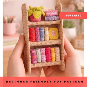 Puede incluir: Una estantería de ganchillo en miniatura llena de libros de ganchillo coloridos, un cuadrado amarillo y una planta. La estantería es beige y tiene tres estantes. El texto "BEGINNER FRIENDLY PDF PATTERN" está en la parte inferior.