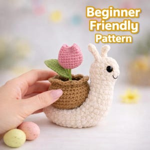 Puede incluir: Un caracol tejido a ganchillo con una maceta en la espalda, con un tulipán rosa y hojas verdes. El caracol es de color blanco roto, con ojos negros y una boca sonriente. El texto "Beginner Friendly Pattern" se muestra.