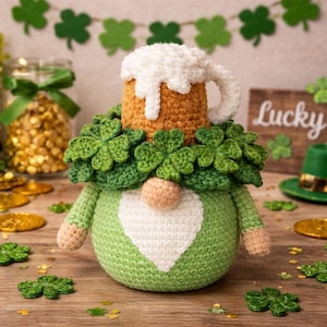May include: A green and white crochet gnome with a shamrock garland and a beer mug hat. The gnome has a peach-colored nose and is decorated with gold coins and shamrock accents. A wooden sign with the word "Lucky" is in the background.