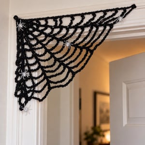 May include: A black crocheted spider web decoration designed to fit into a corner, featuring a series of interconnected yarn loops forming a web pattern. Several small, white plastic spider figures are attached to the black web.