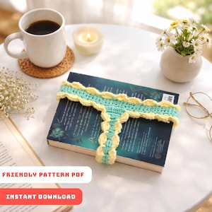 Pode incluir: Um marcador de livros de crochê em azul claro e amarelo com borda ondulada, colocado em um livro. Uma xícara de café, uma vela acesa e um pequeno vaso de flores também estão na mesa. O texto "FRIENDLY PATTERN PDF" e "INSTANT DOWNLOAD" são visíveis.