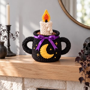 May include: A black crocheted cauldron with two handles, featuring a yellow crescent moon and small star embroidery. A white crocheted candle with a yellow and orange flame sits inside, accented by a purple ribbon tied in a bow.