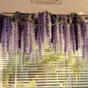 May include: A handcrafted crochet window valance featuring cascading wisteria blooms in shades of purple and white. The valance is adorned with green leaves and hangs above a window with horizontal blinds, creating a decorative floral accent.
