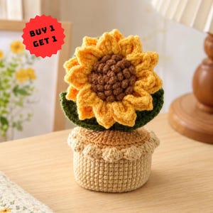 May include: A crocheted sunflower plant in a beige pot with a textured brown centre and bright yellow petals. A dark green leaf sits behind the flower. A red star-shaped graphic in the upper left corner displays the text BUY 1 GET 1.