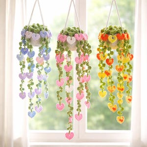 Könnte beinhalten: Drei hängende Häkelpflanzgefäße mit kaskadenförmigen herzförmigen Ranken. Jedes Pflanzgefäß hat ein anderes Farbschema: blau, rosa und gelb. Die Pflanzgefäße sind an weißen Kordeln aufgehängt und schaffen eine charmante, handgefertigte Ästhetik. Die Pflanzgefäße sind mit grünem, strukturiertem Häkelwerk gefüllt.