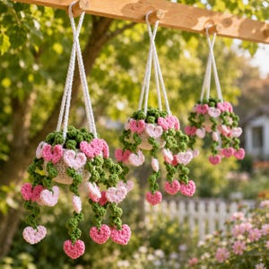 May include: Three hanging crochet plant baskets featuring white pots and trailing green vines adorned with small pink and light pink heart-shaped flowers. The items are suspended by white braided cords from a wooden beam in an outdoor setting.