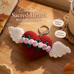 Op de afbeelding: Een rode gehaakte hart-sleutelhanger met witte vleugels, versierd met een band van kleine roze roosjes en groene blaadjes. De tekst luidt: Crochet Sacred Heart Keychain Pattern, Easy to Follow, Instant Download. Inclusief een zilveren metalen ring.
