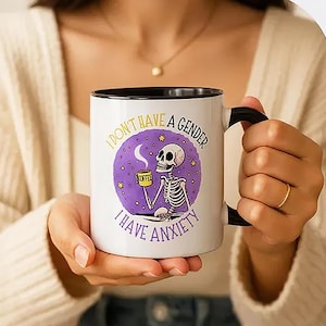 May include: A white ceramic mug with a black handle and rim, featuring a graphic of a skeleton holding a yellow coffee cup. The purple circular background includes stars and the text "I DON'T HAVE A GENDER I HAVE ANXIETY" in black lettering.