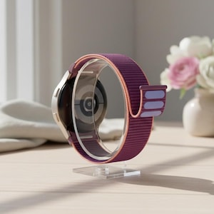 May include: A modern smartwatch with a purple and pink band is displayed on a clear acrylic stand. The watch has a silver case and a black screen. A small vase of flowers sits in the background.