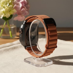 May include: A black smartwatch with a terracotta-coloured fabric strap, displayed on a clear acrylic stand. The watch features a circular dial and a black clasp. The strap is textured and wraps around the watch face.