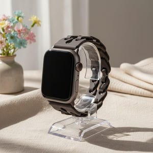 May include: A gray smartwatch with a dark gray chain-link band is displayed on a clear acrylic stand. The watch face is black. A small vase of flowers sits in the background.