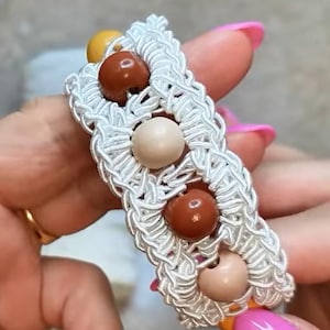 Può includere: Un braccialetto in macramè fatto a mano con un motivo intrecciato bianco. Il braccialetto è adornato da una serie di perline rotonde nei toni del marrone, beige e giallo. Il braccialetto è tenuto da una persona con lo smalto rosa.