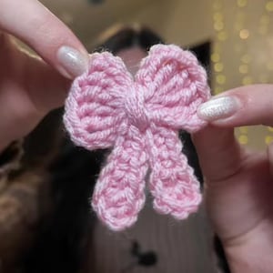 May include: A handmade, light pink crochet bow. The bow is held between two hands, showcasing its delicate, textured design. The bow's loops and center knot are clearly defined, highlighting the craftsmanship. This accessory could be used as a hair clip or decorative element.